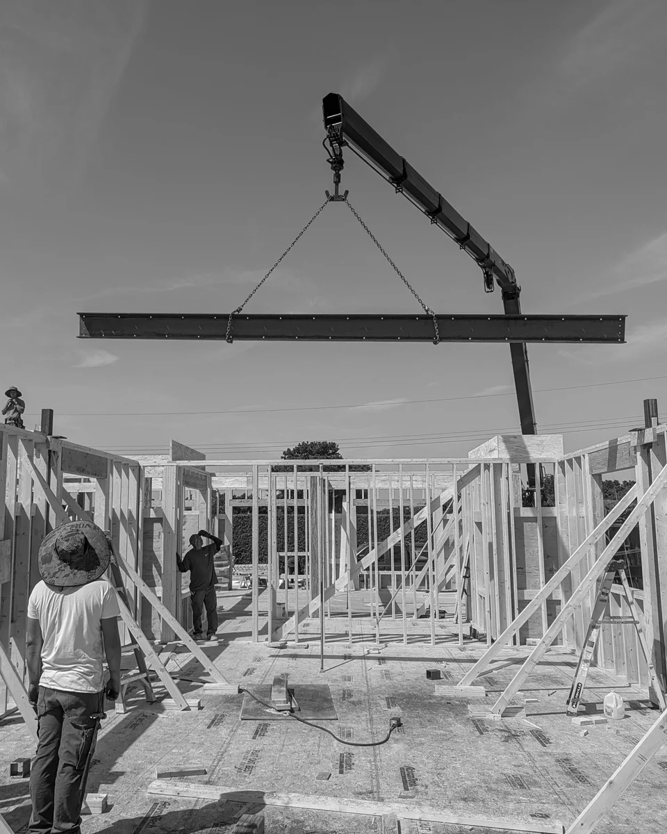 A crane lowers a steel beam toward timber framing while the site crew watches its placement.